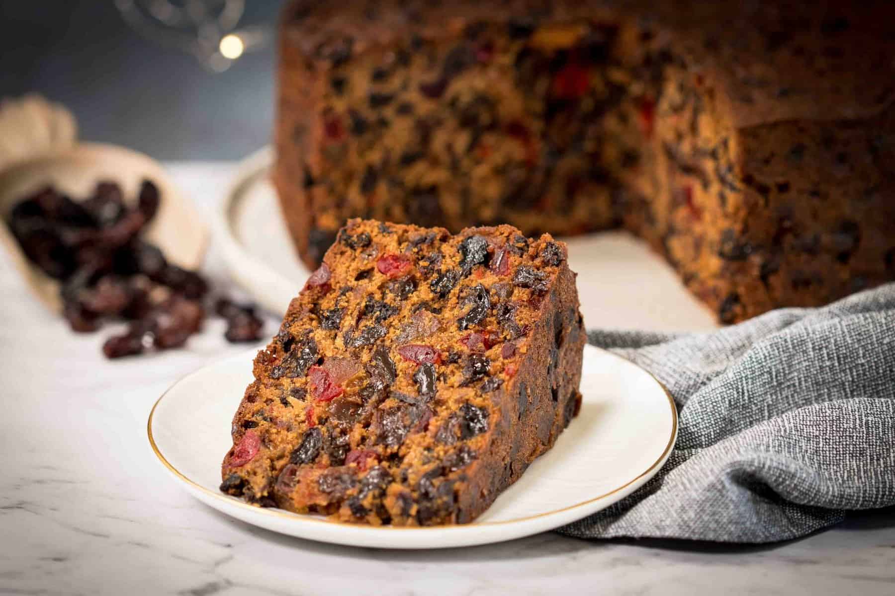 A thick slice of classic Christmas fruitcake on a small plate, showing a dark, moist crumb packed with dried fruit and nuts, with a mug of tea nearby on a warm kitchen table