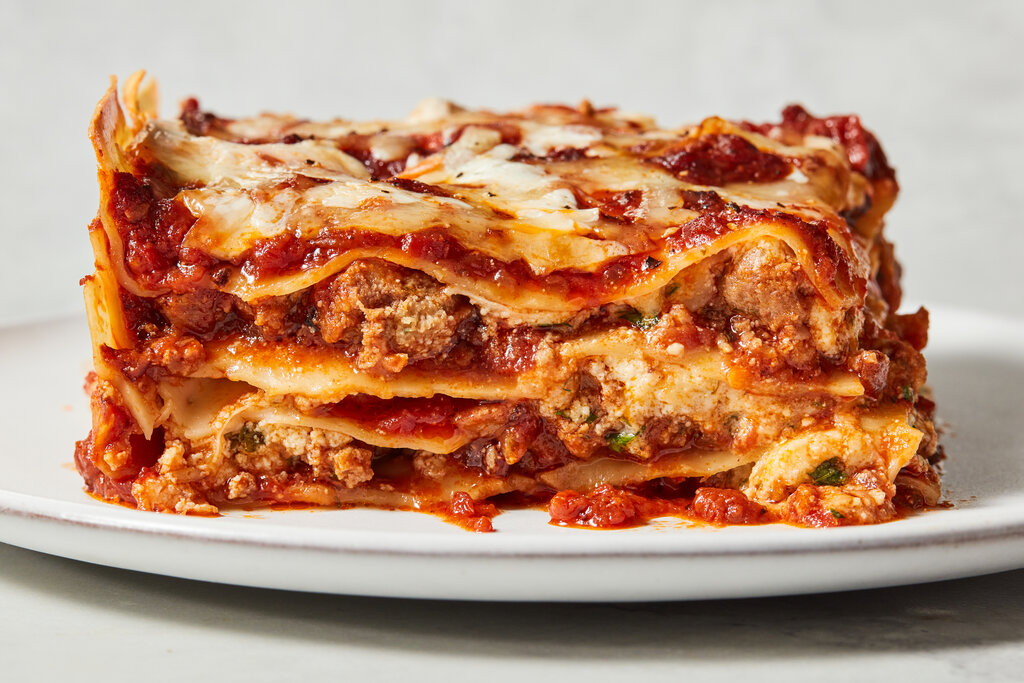 A thick slice of classic meat lasagna on a white plate with melted mozzarella on top and a little extra sauce pooled beside it, warm kitchen table lighting
