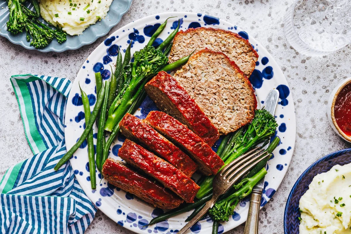 A thick slice of meatloaf with oatmeal and a glossy ketchup glaze on top, served on a white plate with mashed potatoes and green beans on a warmly lit kitchen table