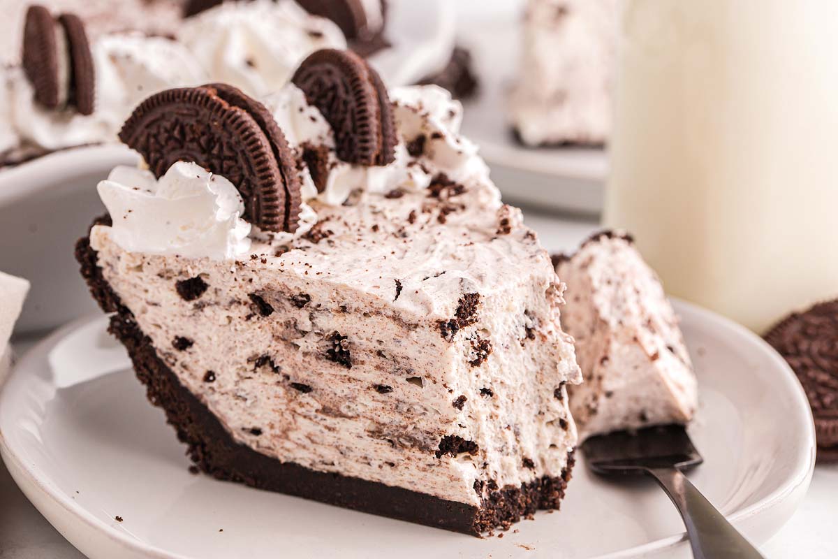 A thick slice of no-bake Oreo pie on a white dessert plate, showing a dark Oreo crumb crust and a creamy cookies-and-cream filling, with a few cookie crumbs scattered on the plate
