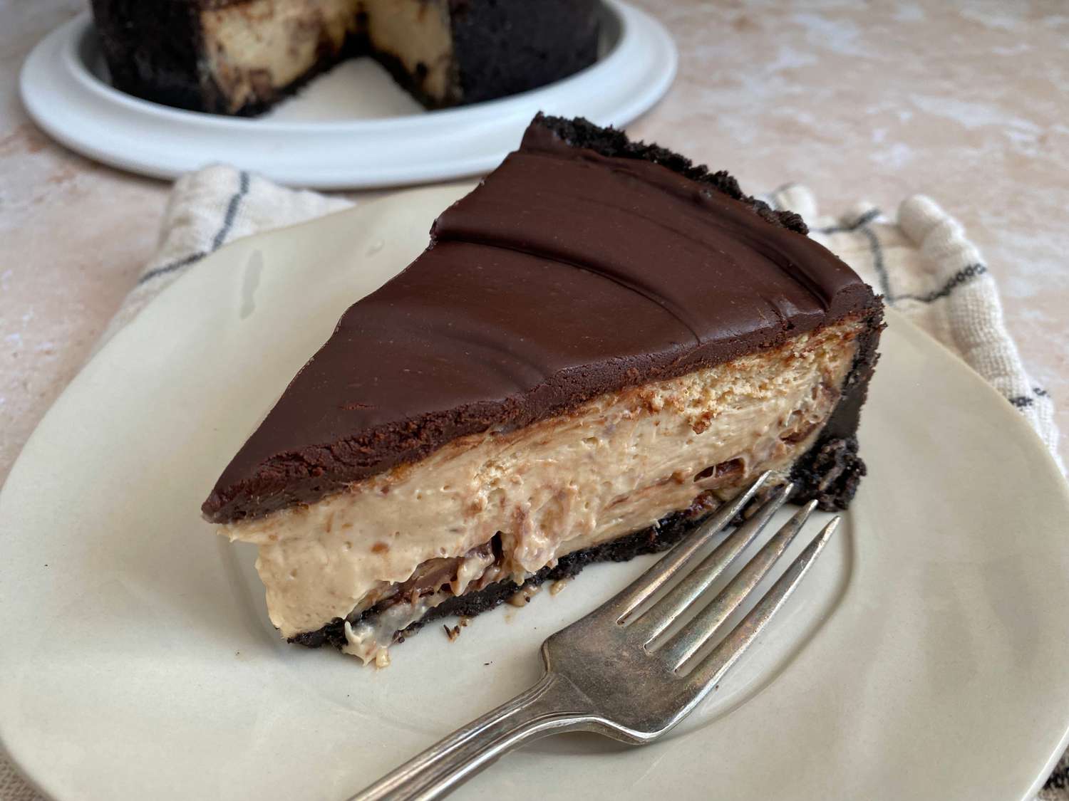 A thick slice of peanut butter cheesecake on a white plate with a glossy chocolate ganache topping and a few chopped peanuts scattered around, set on a warm kitchen table