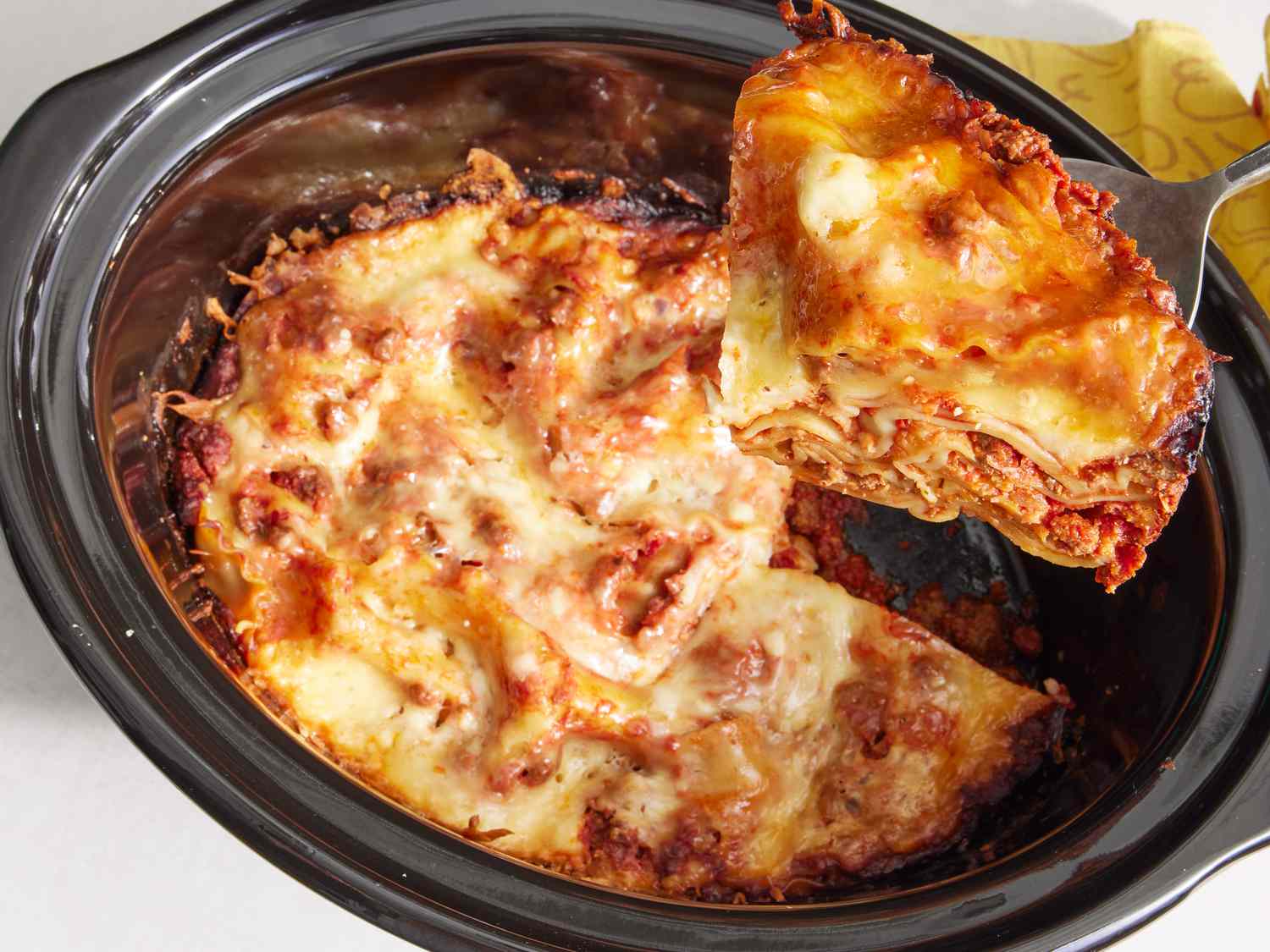 A thick slice of slow cooker lasagna on a white plate with melted mozzarella on top, a little sauce pooling at the edge, and a fork resting beside it on a warm kitchen table