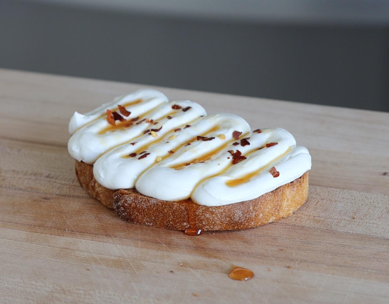 A thick slice of toasted sourdough topped with fluffy whipped ricotta, drizzled with hot honey, finished with lemon zest and chopped herbs on a sunlit kitchen counter