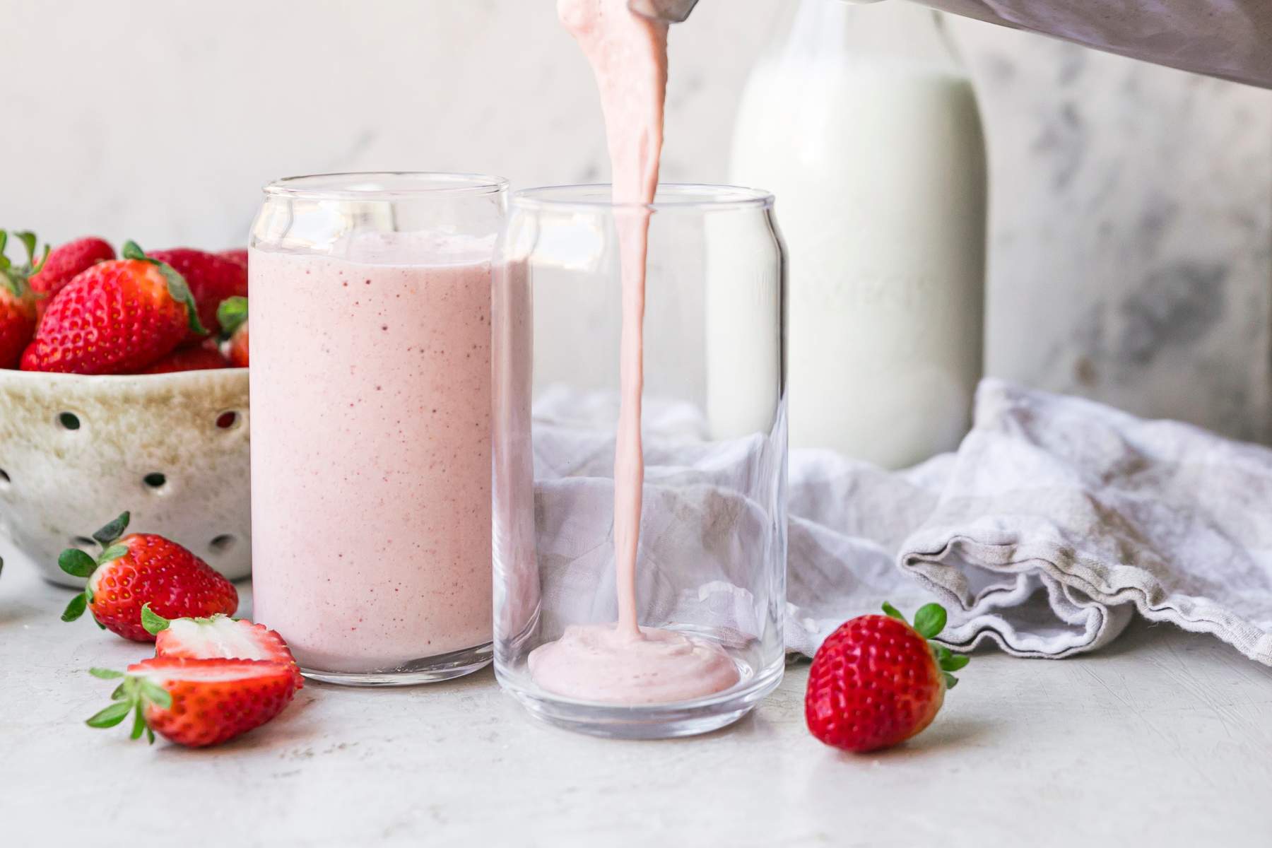 A thick strawberry milkshake being poured from a blender into a chilled glass on a clean kitchen counter with soft natural light