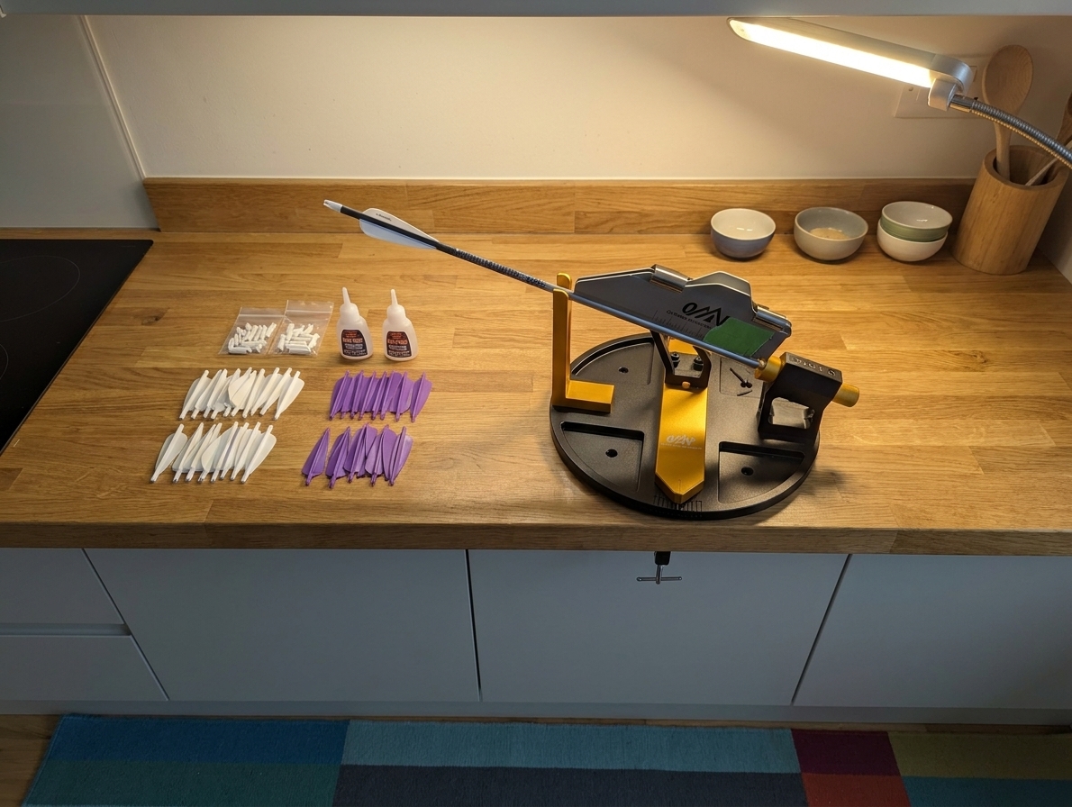 A tidy kitchen counter set up as a fletching table with an arrow fletching jig, vanes, nocks, small bottles of adhesive, and a bright task light on a clean, flat surface