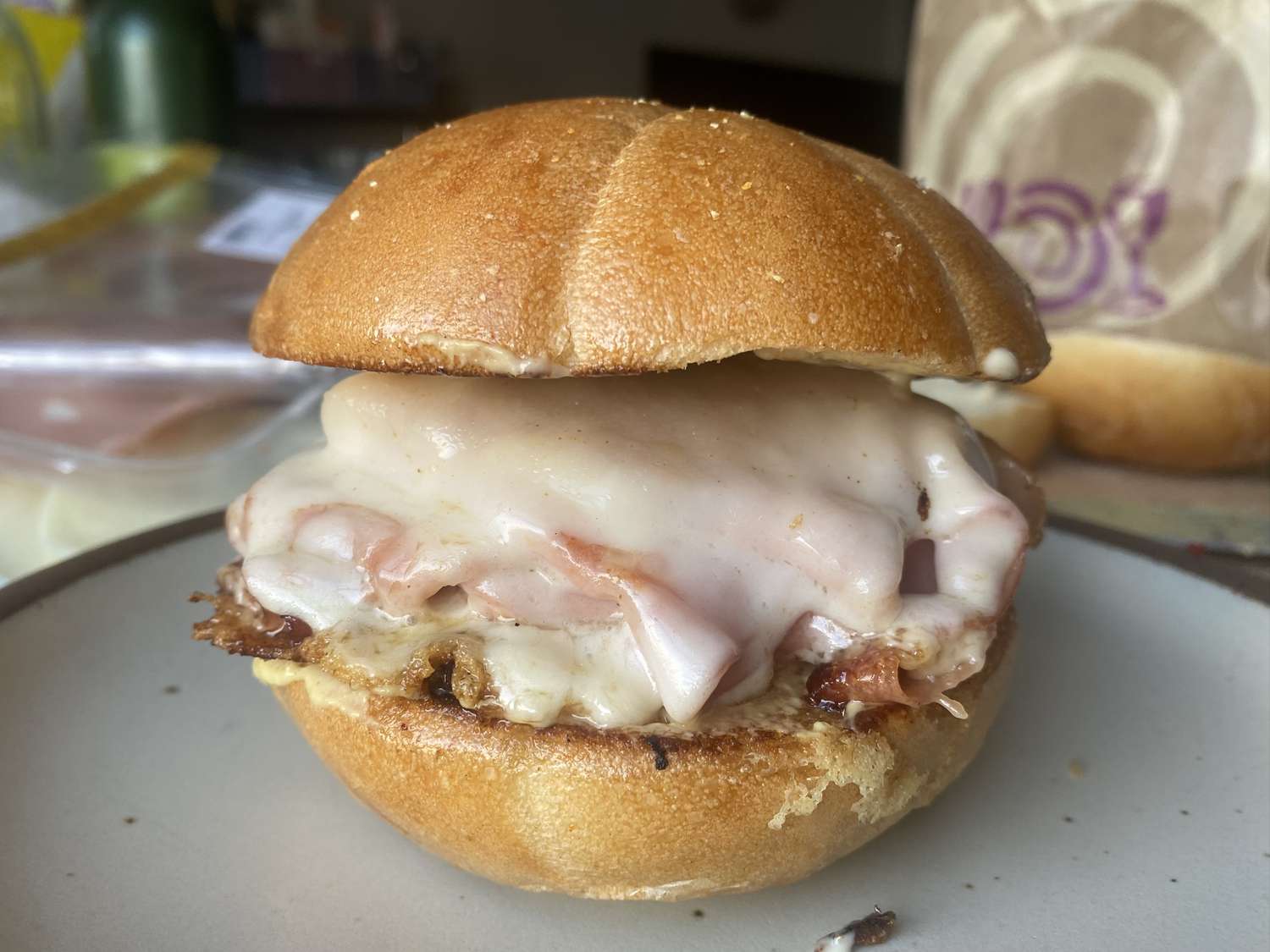 A toasted roll sandwich stuffed with crispy-edged mortadella and melted provolone on a wooden cutting board with a small dish of mustard nearby