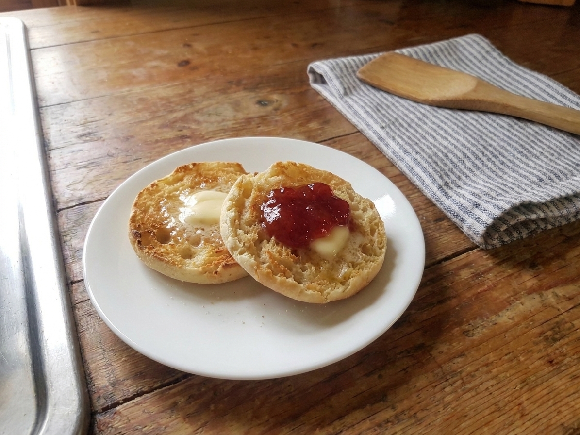 A toasted split muffin on a plate with melting butter and a spoonful of strawberry jam, set on a breakfast table