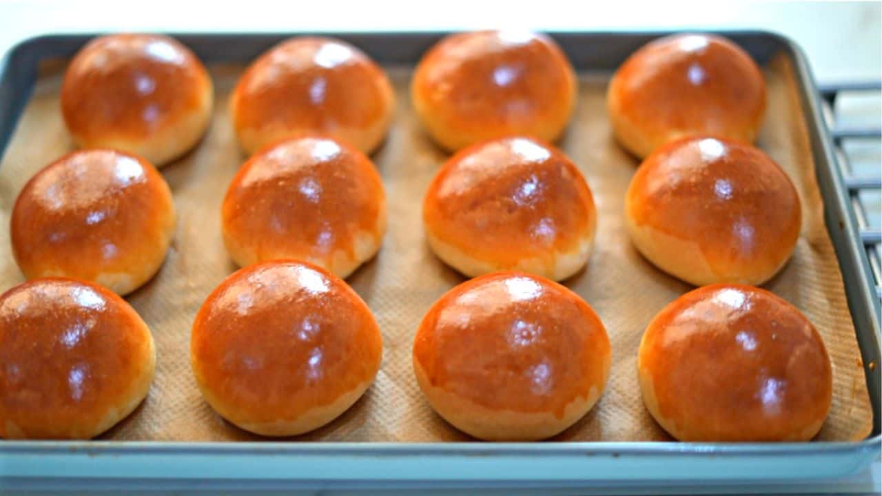 A tray of freshly baked brioche buns with glossy golden tops cooling on a parchment-lined baking sheet on a bright kitchen counter