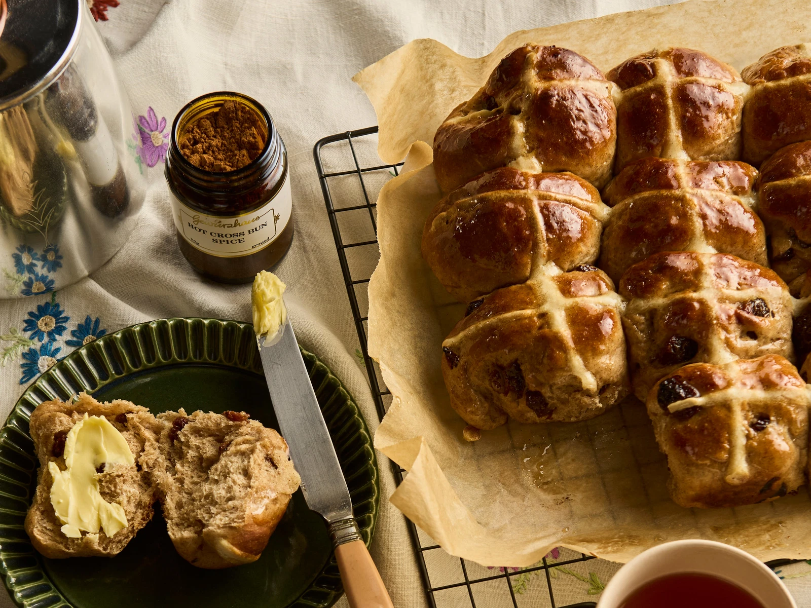 A tray of freshly baked hot cross buns with shiny glazed tops and piped white crosses, cooling on a parchment-lined baking sheet on a bright kitchen counter