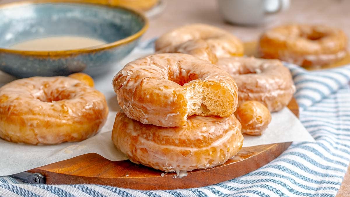 A tray of freshly glazed yeast donuts with a glossy vanilla sheen on a parchment-lined baking sheet