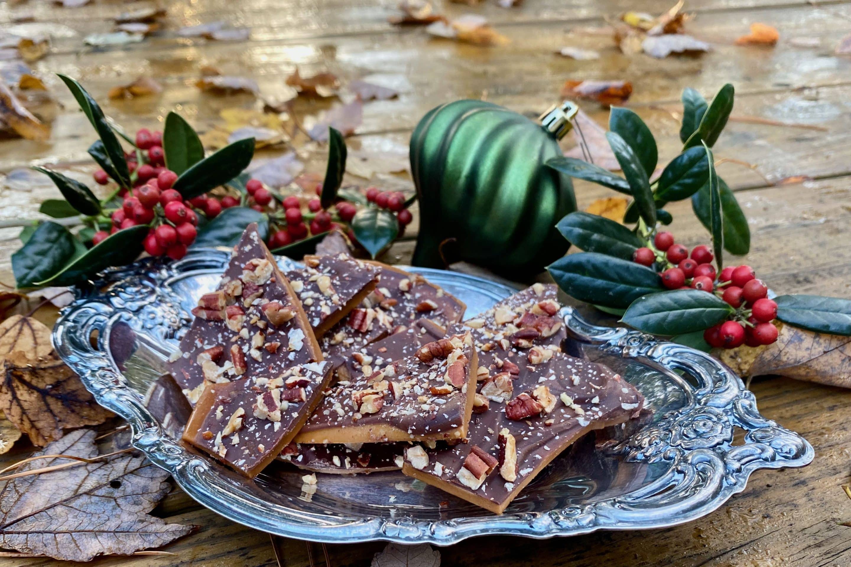 A tray of glossy chocolate-topped Christmas toffee broken into rustic pieces on parchment paper, with toasted nuts scattered around on a warm kitchen counter