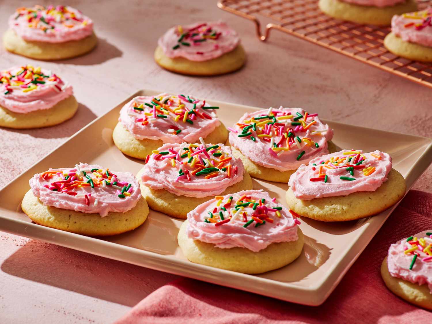 A tray of soft, thick sugar cookies topped with pale pink frosting and rainbow sprinkles on a light stone kitchen counter with warm natural light
