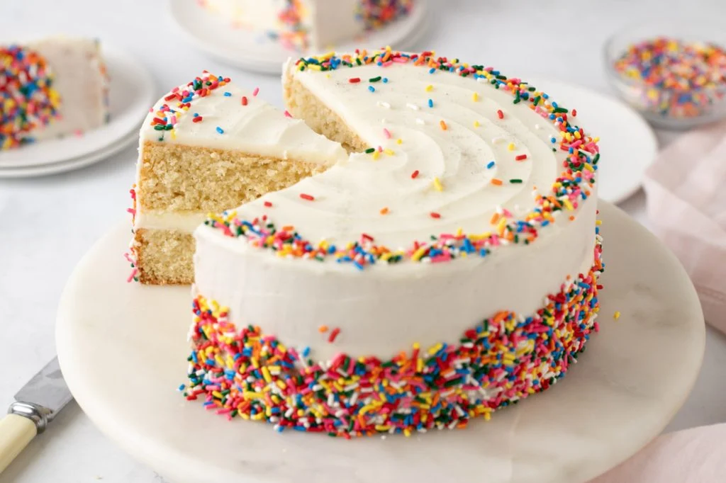 A two-layer gluten free vanilla cake with vanilla buttercream frosting on a white cake stand on a bright kitchen counter, natural window light