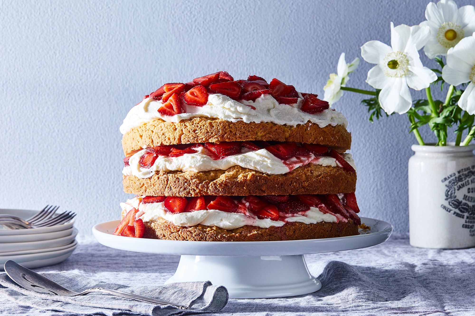 A two layer strawberry shortcake cake frosted with whipped cream and topped with fresh strawberries on a simple white cake stand