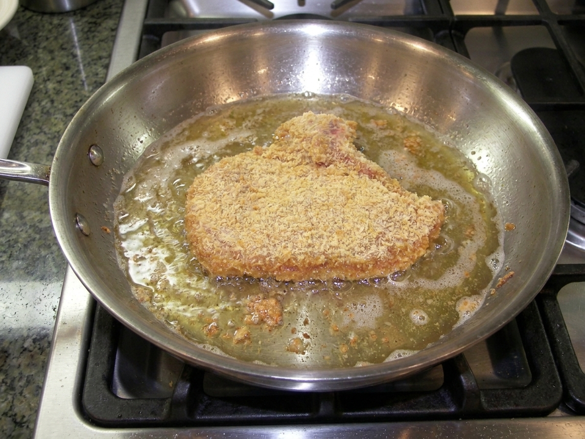 A veal cutlet frying in bubbling oil in a stainless steel skillet on a stovetop, turning golden brown around the edges