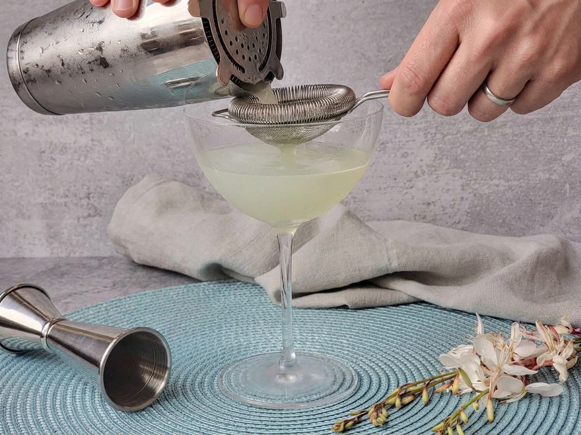 A vodka gimlet being strained through a cocktail strainer into a chilled coupe glass on a stone countertop