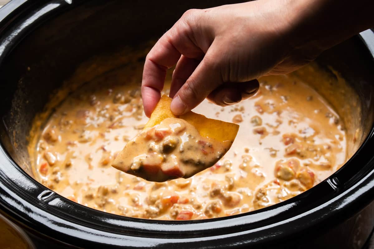 A warm bowl of creamy Velveeta Rotel cheese dip on a wooden table surrounded by tortilla chips and sliced jalapeños, cozy kitchen background