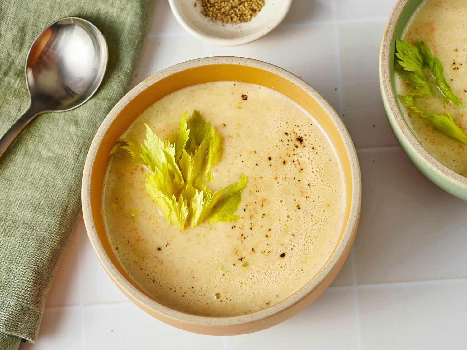 A warm bowl of creamy celery soup garnished with cracked black pepper, chopped parsley, and a drizzle of olive oil on a light wooden table