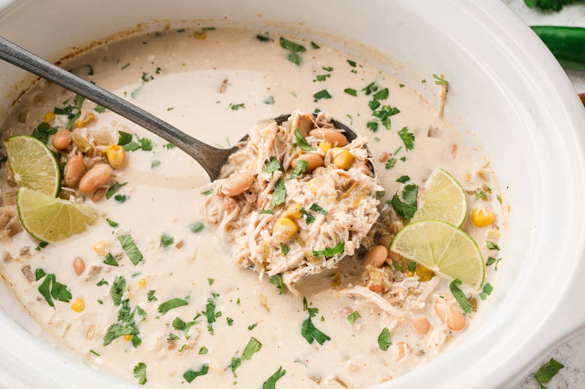 A warm bowl of creamy white chicken chili topped with cilantro, sliced jalapenos, and shredded Monterey Jack on a rustic kitchen table with tortilla chips nearby