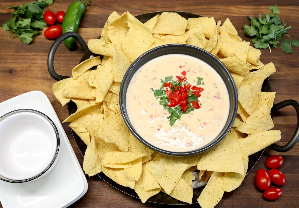 A warm bowl of creamy white queso on a table setting with tortilla chips, salsa <!-- inpage-recommendation:21462 -->, and a small plate of tacos in the background under soft kitchen lighting