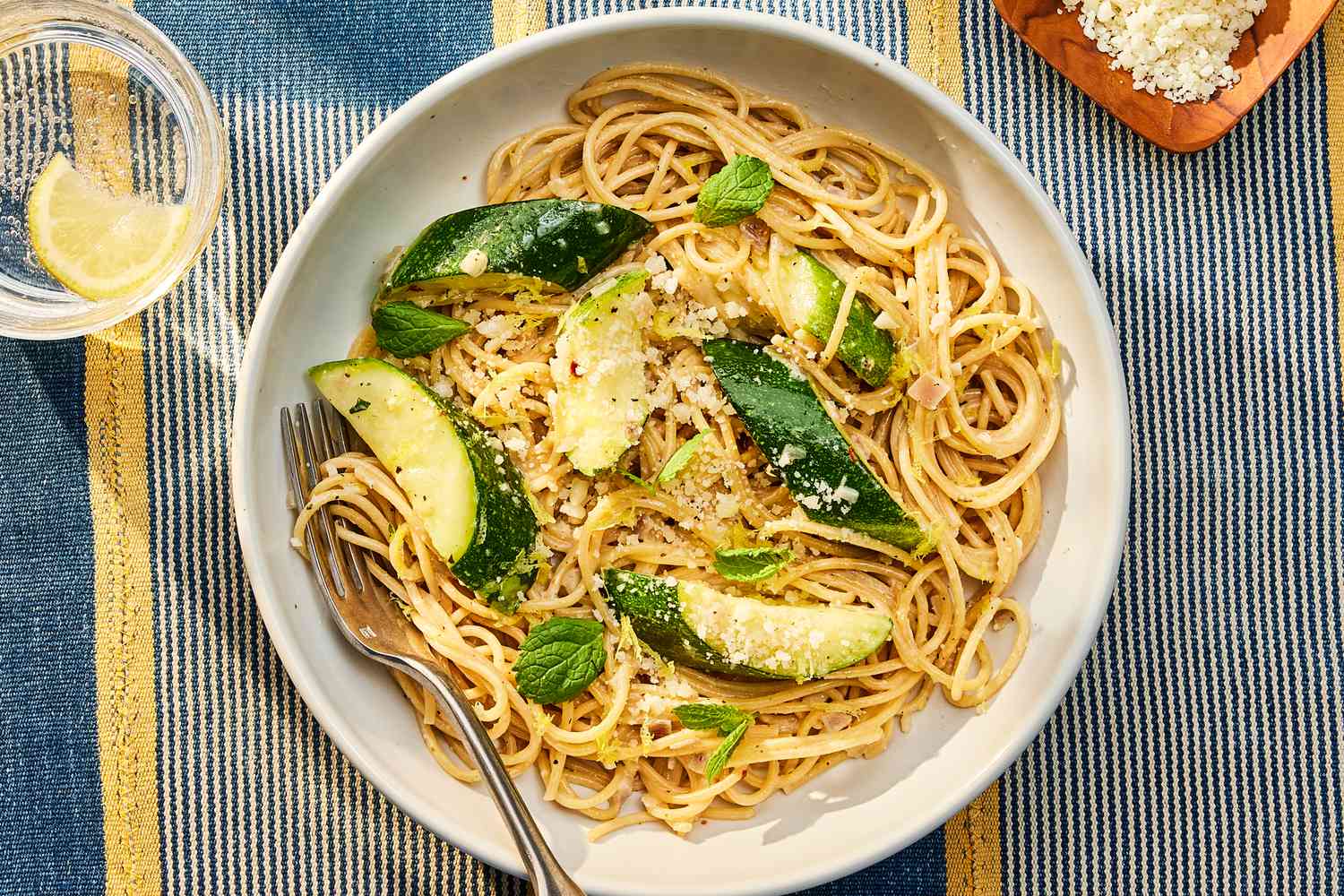 A warm bowl of spaghetti tossed with sautéed zucchini coins, garlic, lemon zest, and grated Parmesan on a light stoneware plate on a wooden kitchen table with soft natural light
