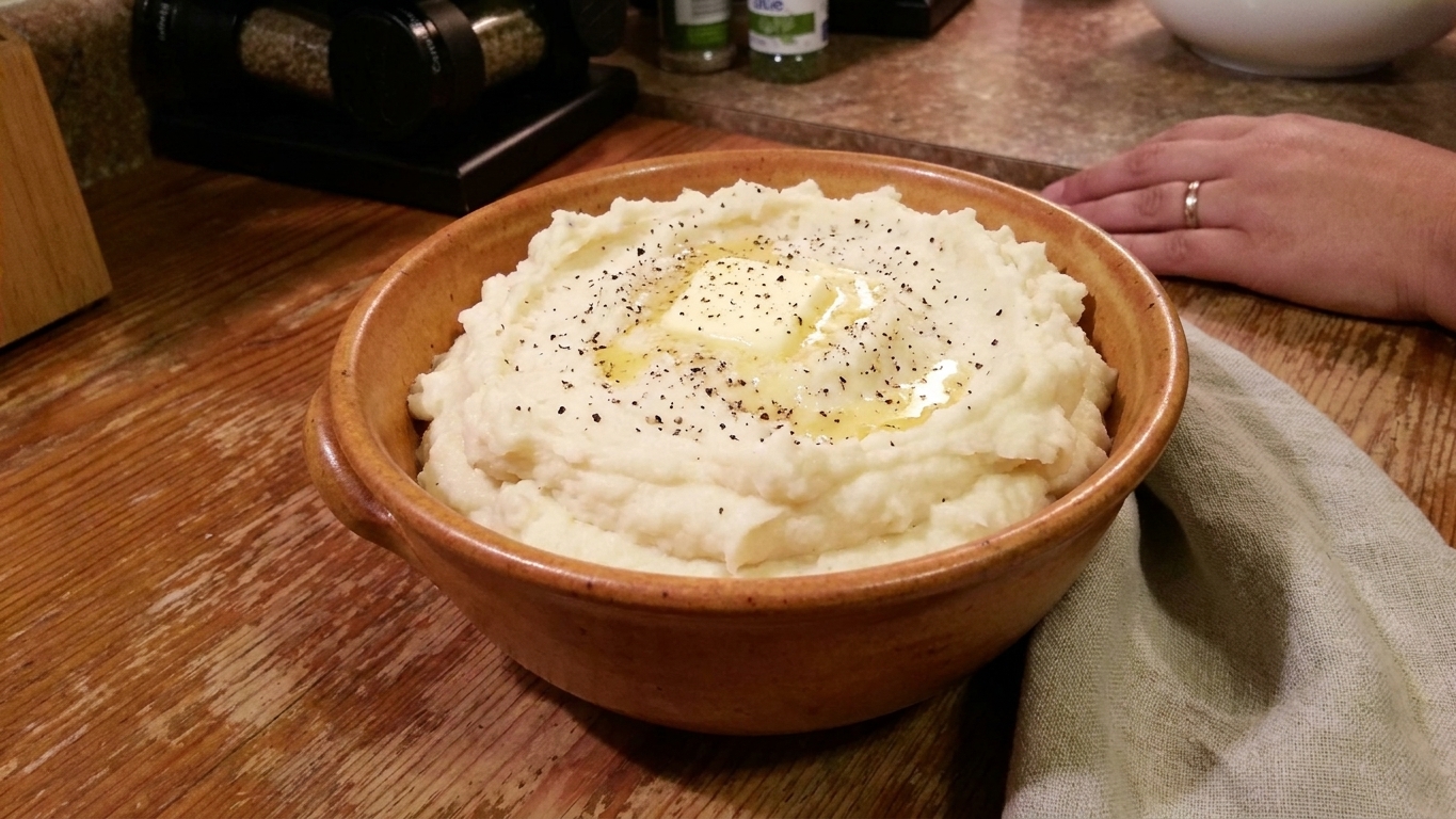 A warm ceramic bowl filled with creamy whipped potatoes topped with a melting pat of butter and cracked black pepper on a wooden table with a linen napkin