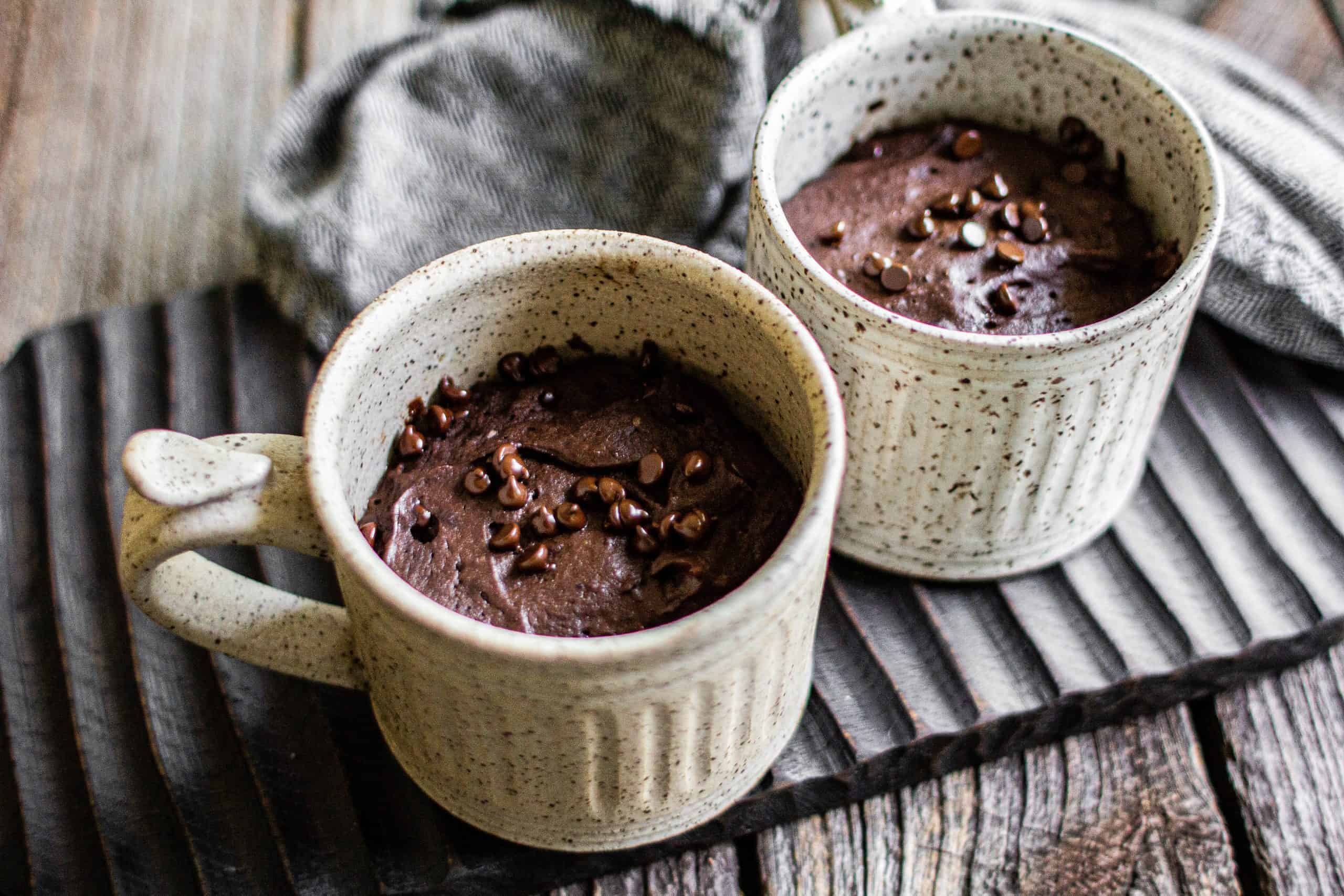 A warm chocolate mug cake in a ceramic mug on a kitchen counter with a spoon beside it, slightly glossy fudgy top and a few chocolate chips melted on the surface