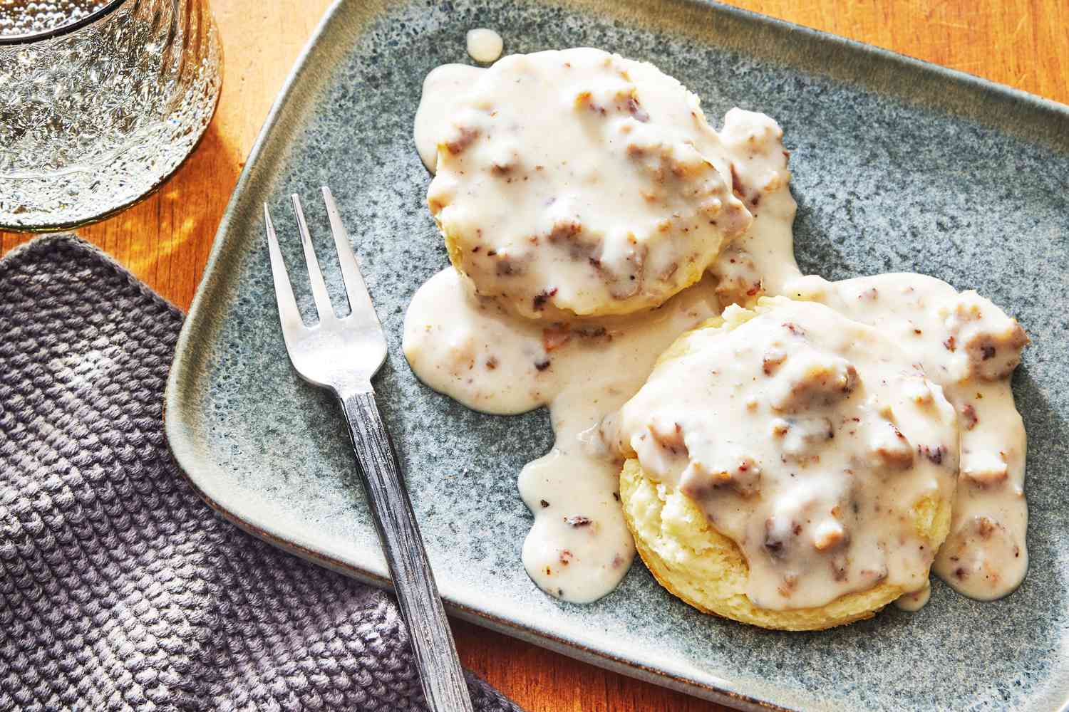 A warm plate of split buttermilk biscuits covered in creamy sausage gravy with lots of black pepper, set on a cozy kitchen table with a simple linen napkin