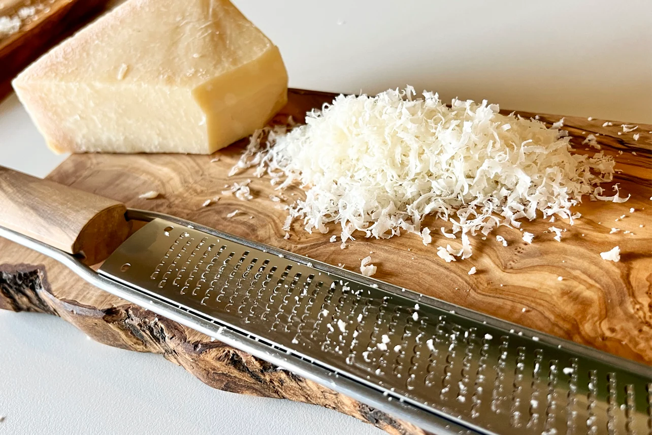 A wedge of Parmesan being finely grated on a microplane into a bowl on a wooden kitchen counter in warm natural light