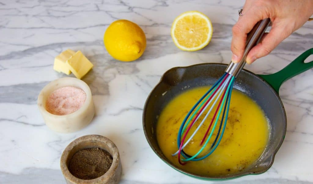 A whisk re-emulsifying glossy lemon butter sauce in a skillet with browned bits, garlic, and herbs on the stovetop
