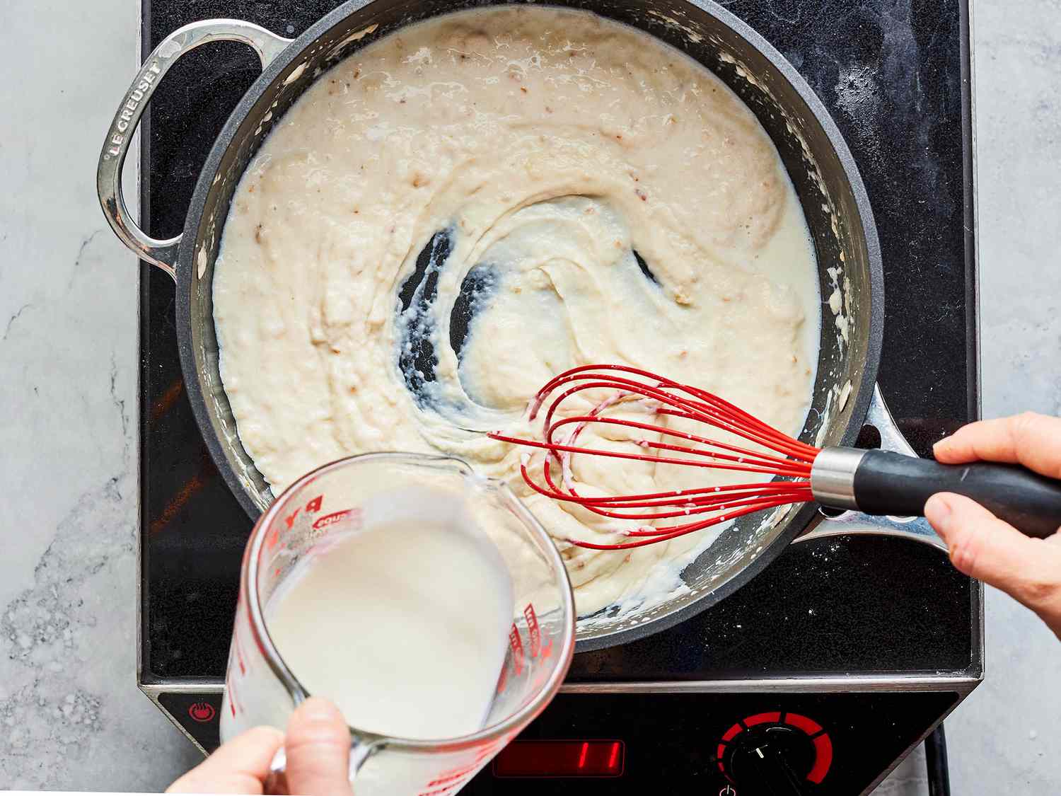 A whisk stirring a creamy sauce in a skillet with sautéed mushrooms and onions, with gentle steam rising from the pan on a stovetop