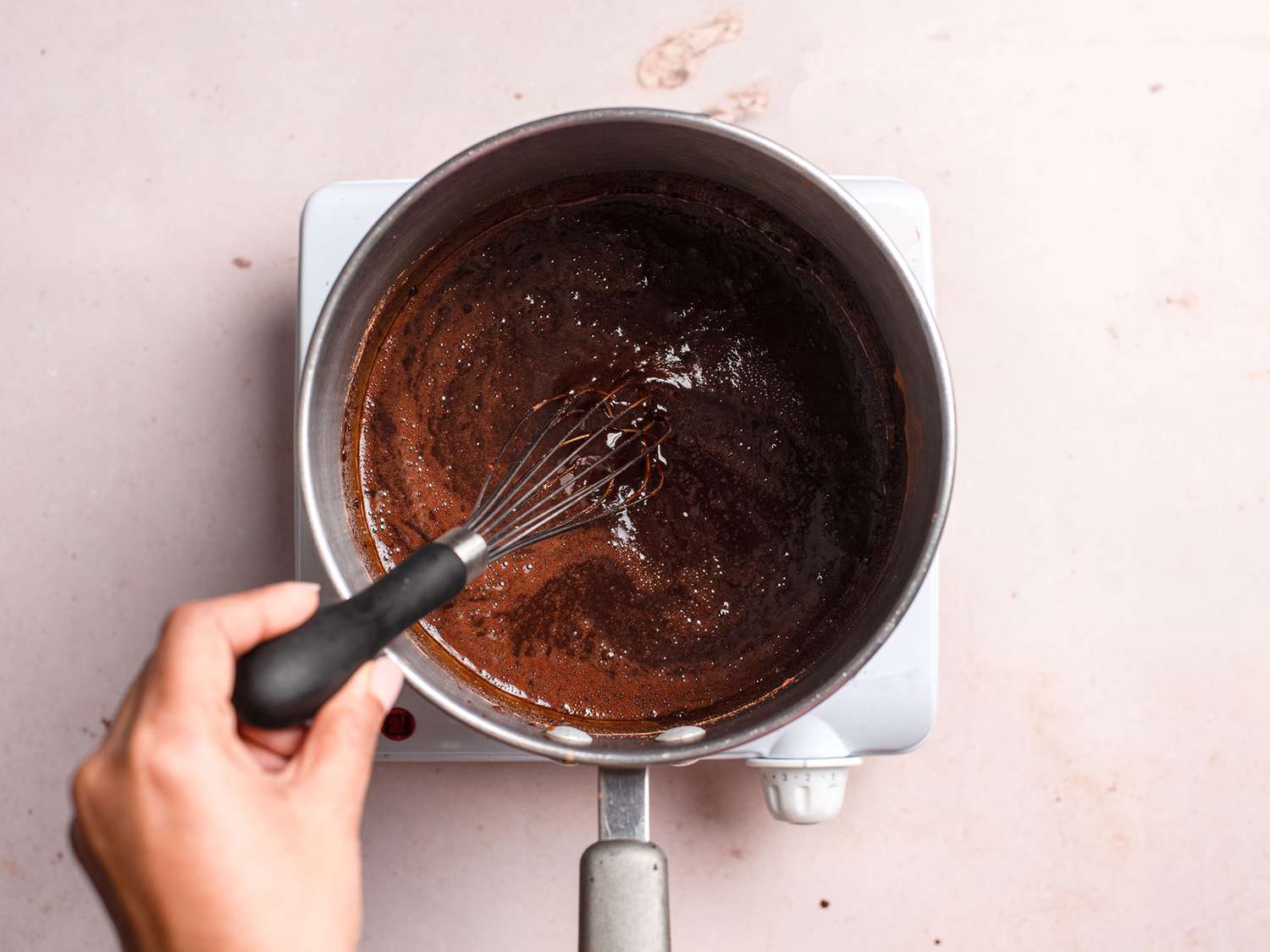 A whisk stirring a thick, smooth cocoa paste in a small saucepan on the stovetop before the milk is added