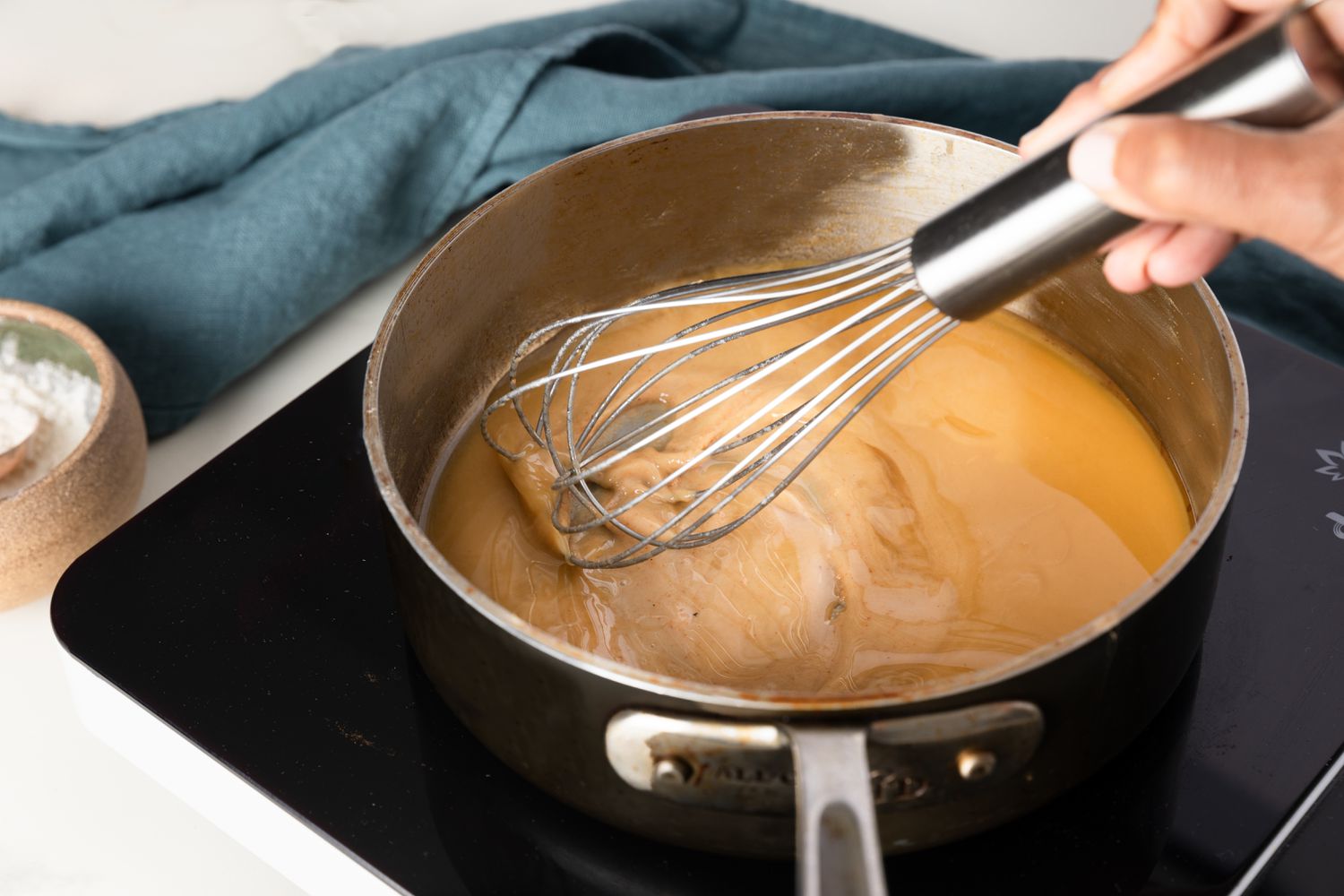 A whisk stirring a thickened milk-and-flour paste in a small saucepan on a stovetop with light steam rising