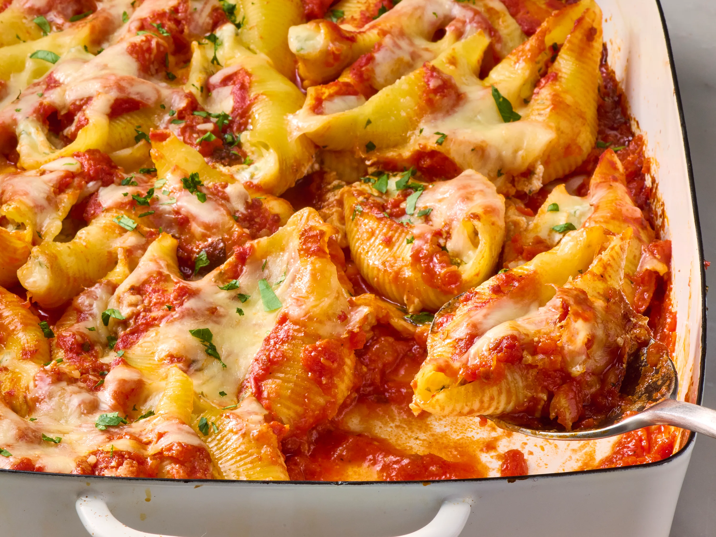 A white baking dish of stuffed pasta shells filled with meat and ricotta, covered in marinara and melted mozzarella, set on a warm kitchen table with a serving spoon