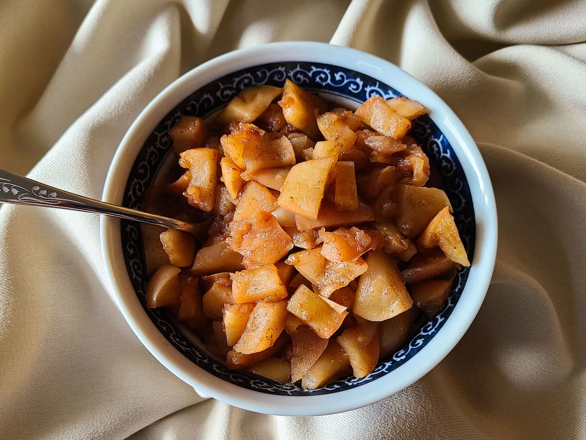 A white bowl filled with glossy cinnamon stewed apples on a light stone countertop, with a spoon and a folded linen towel nearby, warm natural kitchen light