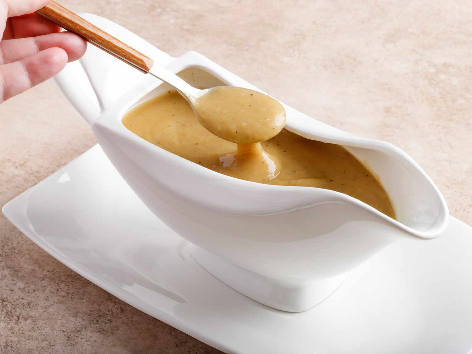 A white bowl filled with glossy, pepper-speckled brown gravy on a wooden table with a spoon resting beside it, warm kitchen lighting