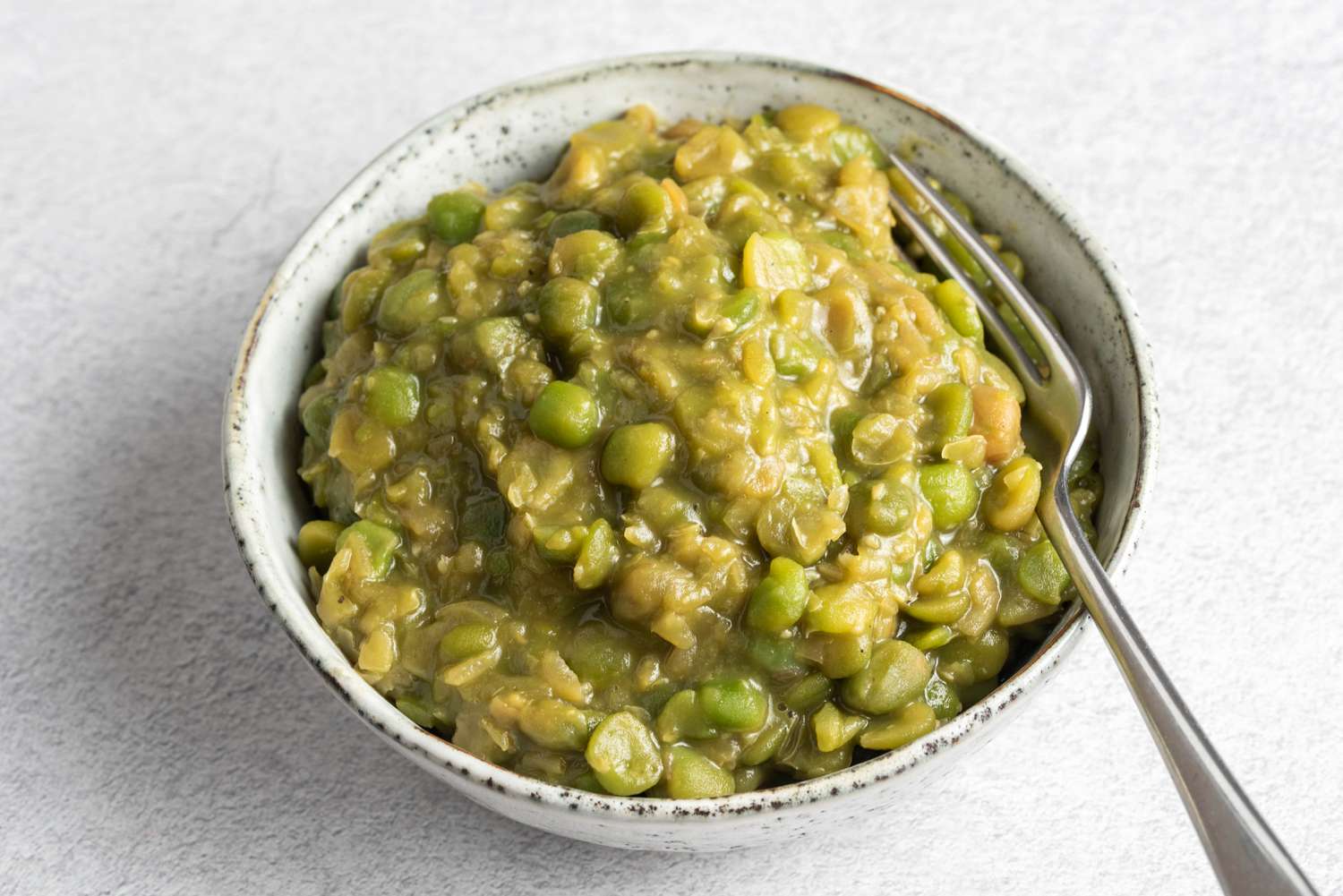 A white bowl of bright green mushy peas with a small pat of melting butter on top, set on a wooden table with a fork and lemon wedge nearby