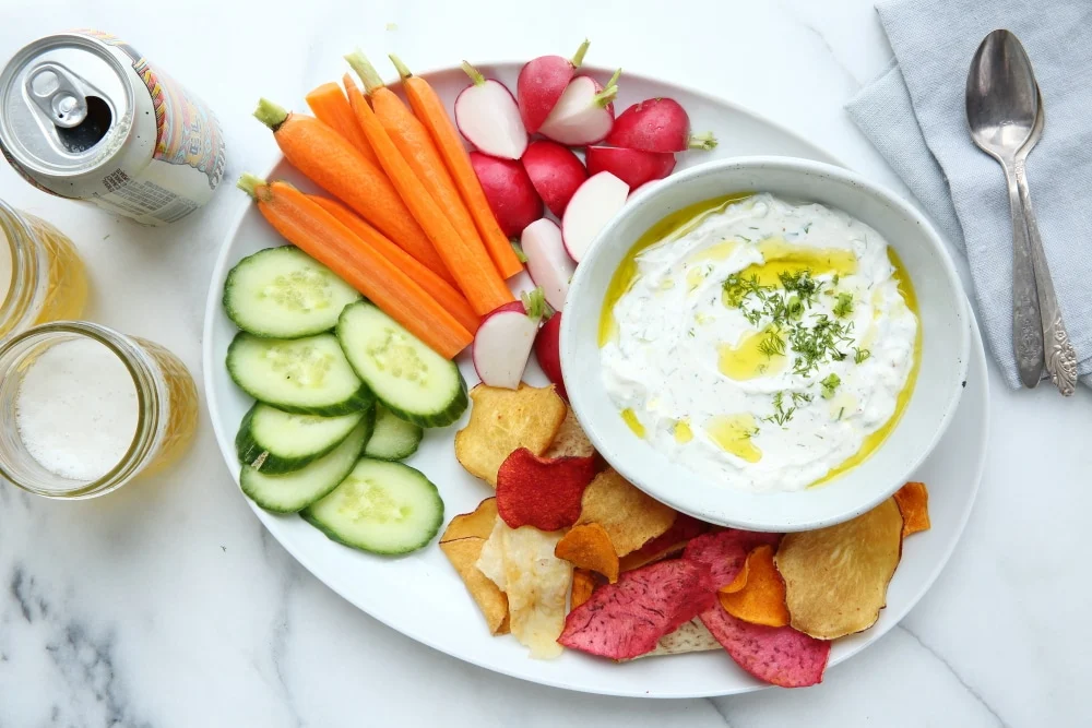 A white bowl of creamy Greek yogurt veggie dip topped with chopped chives and black pepper, surrounded by carrots, cucumbers, and bell pepper strips on a light kitchen counter