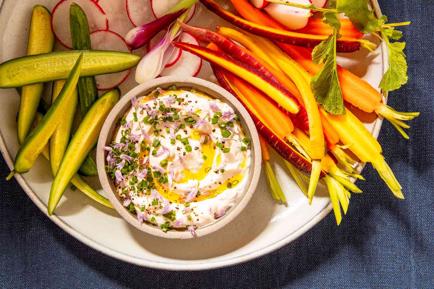A white bowl of creamy herbed vegetable dip on a light kitchen counter, surrounded by carrots, cucumbers, bell pepper strips, and cherry tomatoes with a small spoon