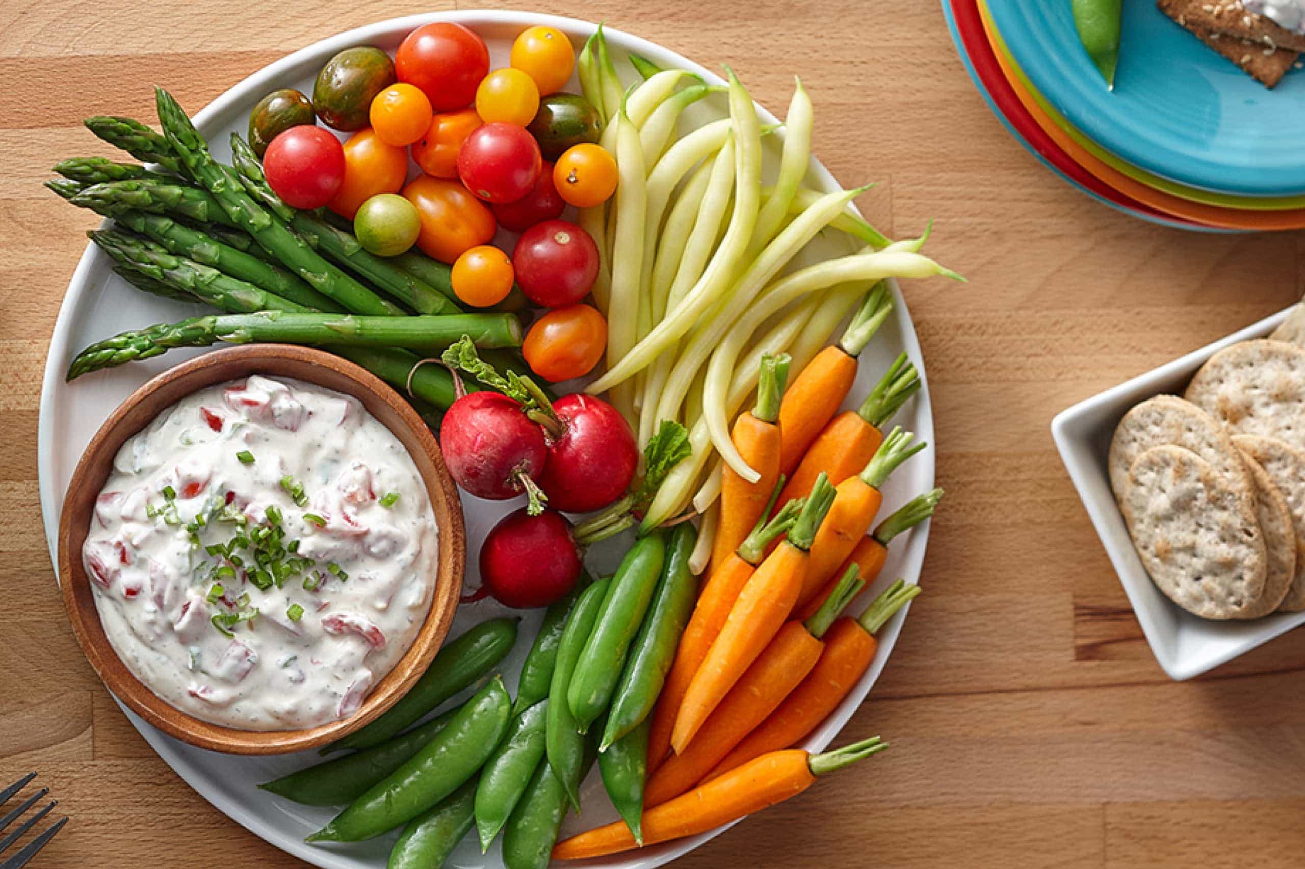 A white bowl of creamy ranch dip topped with chopped chives on a warm wood table, surrounded by carrot sticks, celery, and ridged potato chips