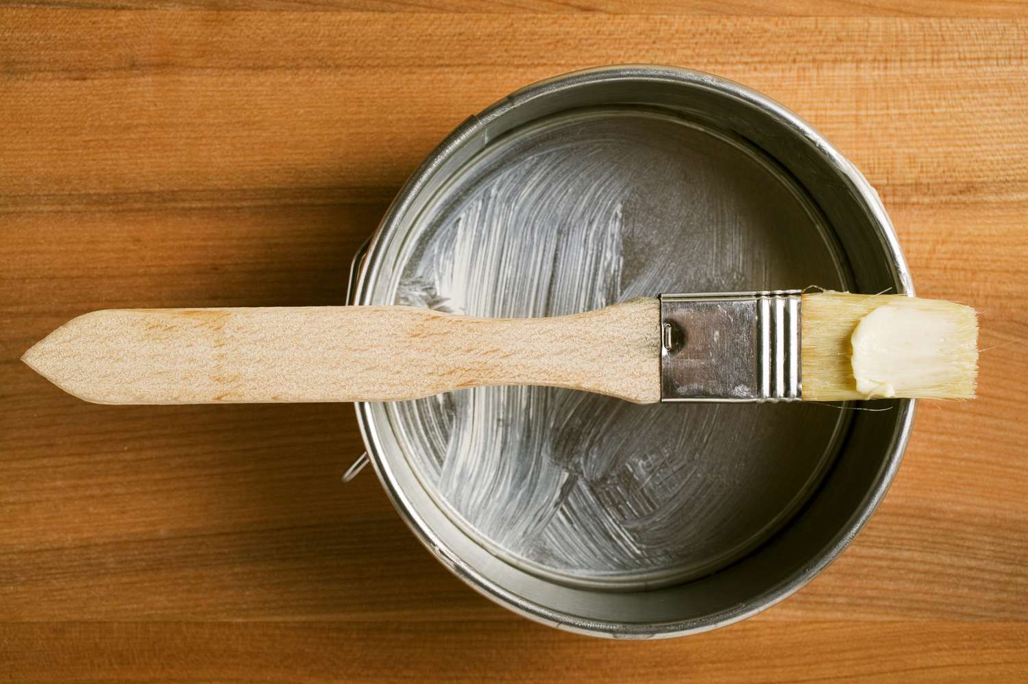 A white ceramic baking dish being lightly buttered on a kitchen counter with soft natural light