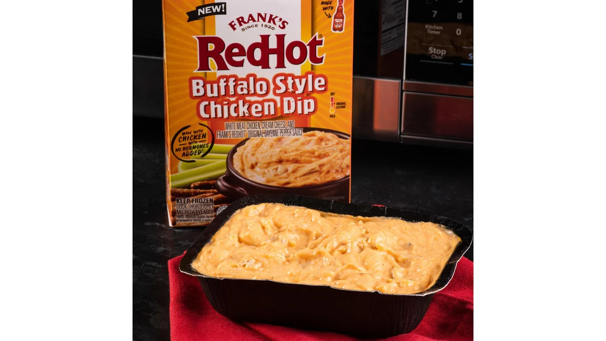 A white ceramic baking dish filled with buffalo chicken wing dip, topped with melted cheddar and green onions, sitting on a warm-toned kitchen counter with tortilla chips nearby