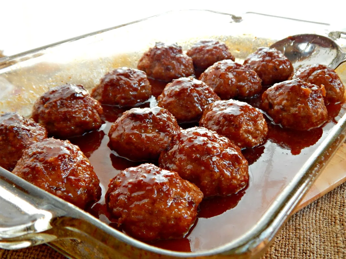 A white ceramic baking dish filled with glossy glazed ham balls, bubbling sauce around them, on a warm kitchen counter with soft natural light