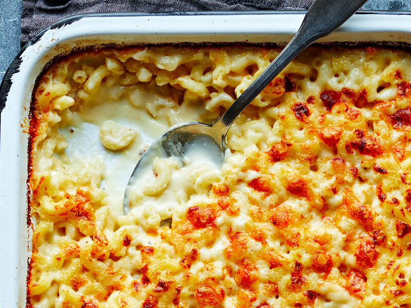 A white ceramic bowl filled with creamy mac and cheese, topped with toasted buttery breadcrumbs, sitting on a warm wood table with a fork beside it