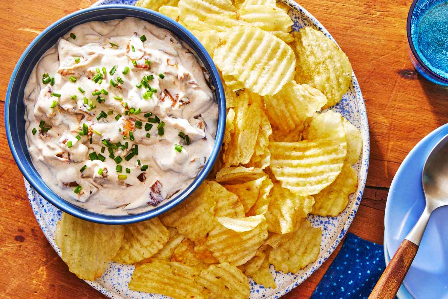 A white ceramic bowl filled with creamy onion chip dip, topped with sliced chives, surrounded by ridged potato chips on a neutral stone countertop in soft natural light