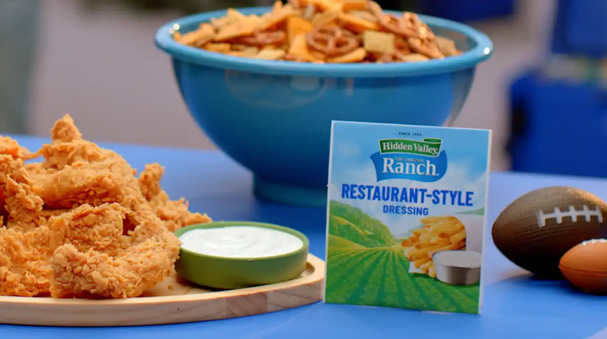 A white ceramic bowl filled with creamy ranch dip on a wooden kitchen counter, surrounded by carrot sticks, celery, and ridged potato chips in warm natural light