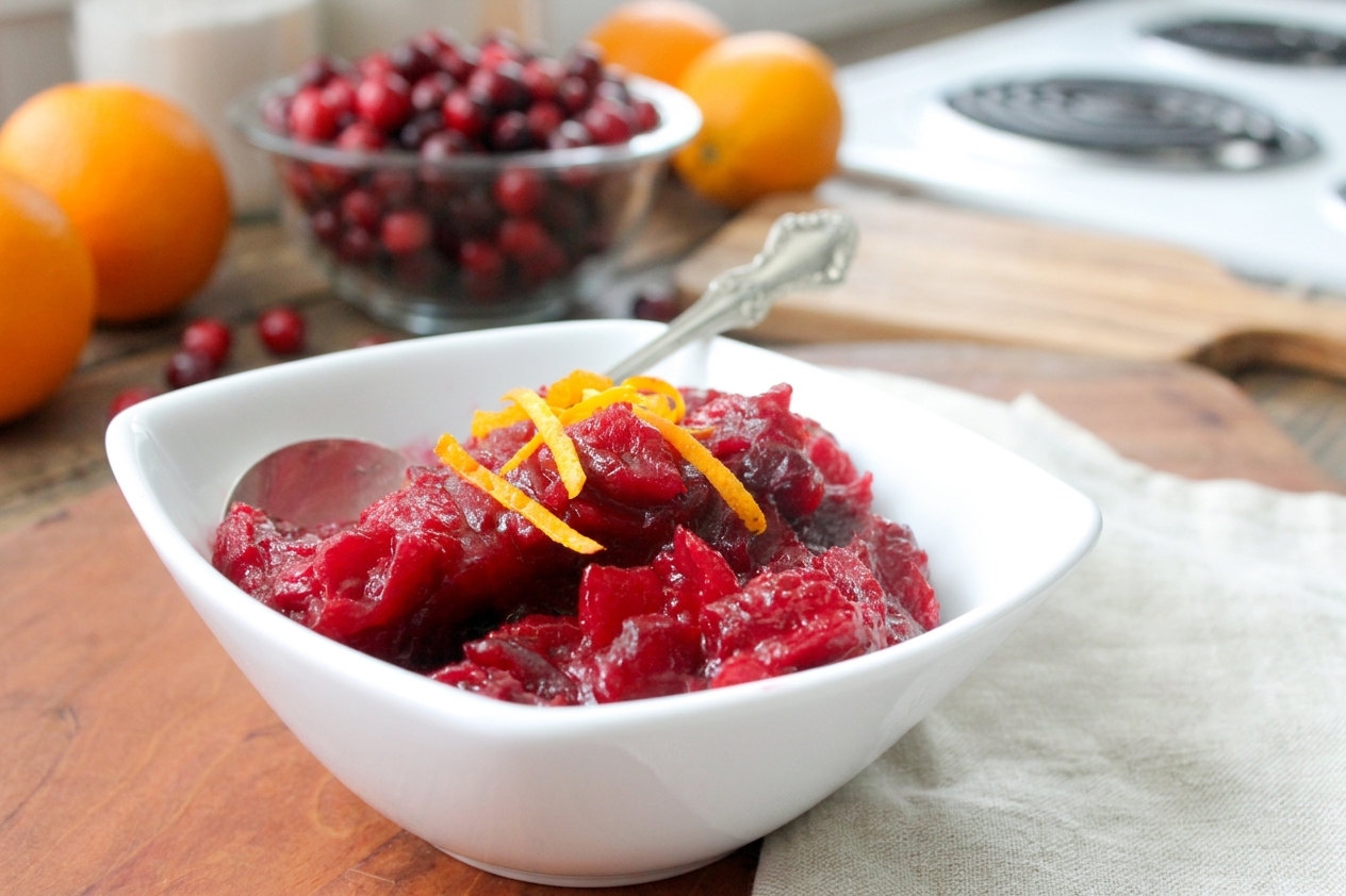 A white ceramic bowl filled with glossy cranberry orange sauce, with visible orange zest and a serving spoon on a cozy kitchen table