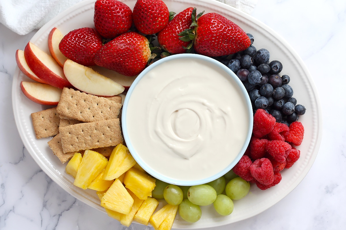 A white ceramic bowl of fluffy cream cheese fruit dip topped with lemon zest on a bright kitchen counter, surrounded by strawberries, grapes, and apple slices