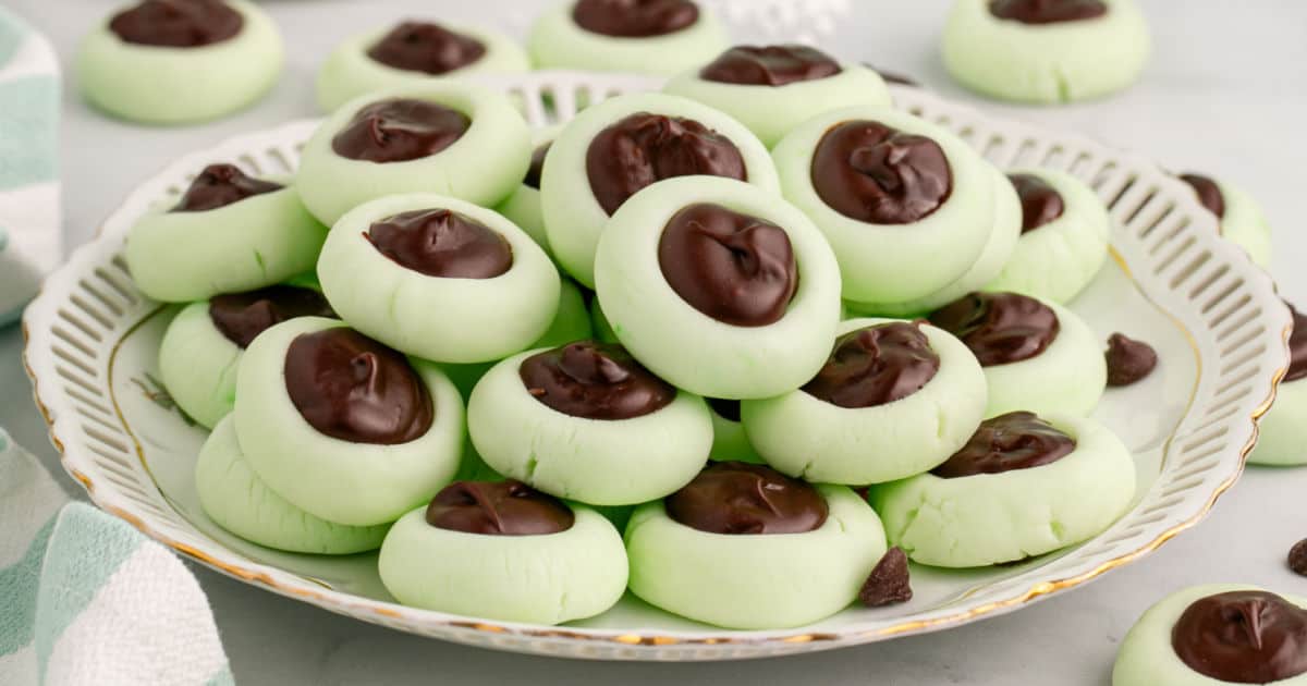 A white ceramic platter piled with pastel cream cheese mints, some pressed with a fork and others shaped into small coins, on a light kitchen table with soft natural light