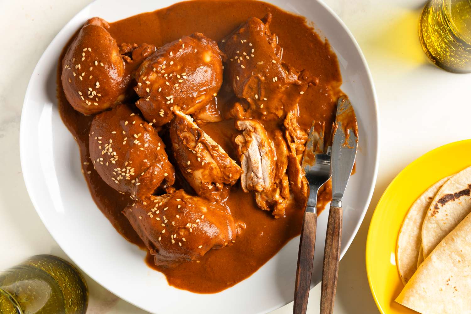A white plate of chicken covered in dark, glossy mole sauce with sesame seeds, served with warm tortillas on a wooden table in soft natural light